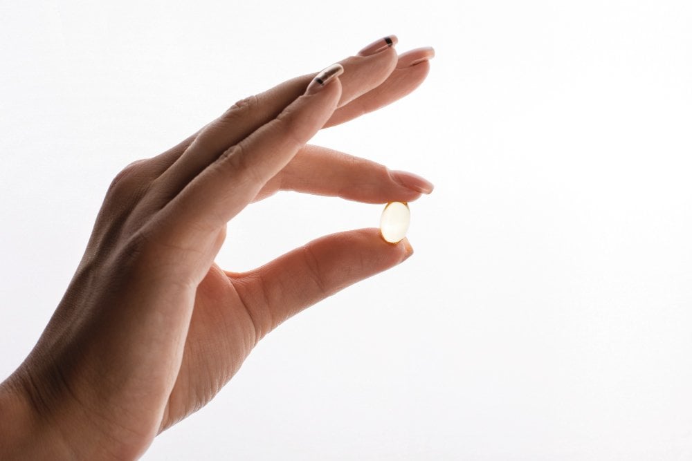 Dłoń trzyma kapsułkę omega-3 między kciukiem a palcem wskazującym na jasnym tle