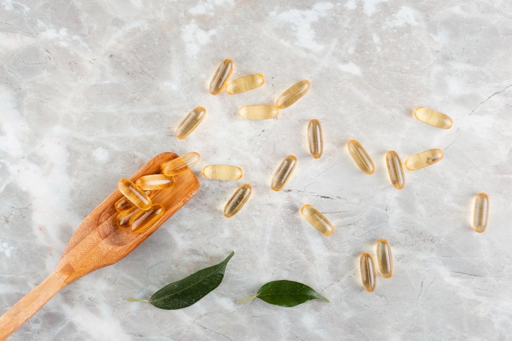 Kapsułki omega-3 na jasnym marmurze, część na drewnianej łyżce, obok dwa zielone liście